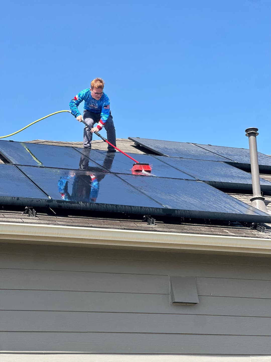 Person cleaning solar panels on a roof with a brush and hose under a clear blue sky.