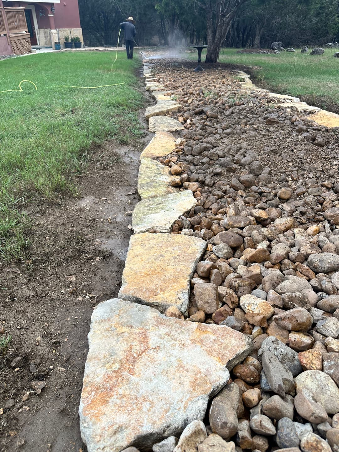 Stone path lined with gravel and large stones in a green yard, with a person watering nearby.