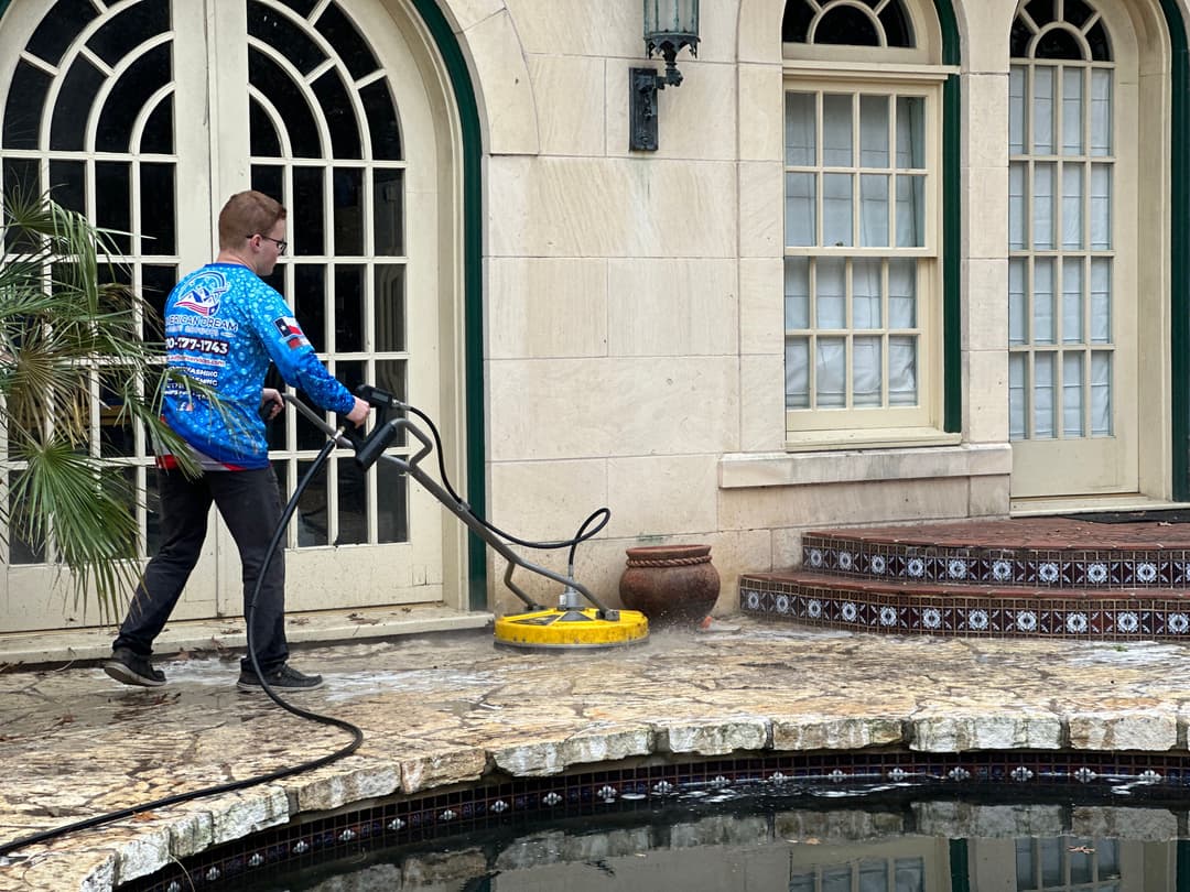Person pressure washing a stone patio near a pool with a yellow cleaning attachment.
